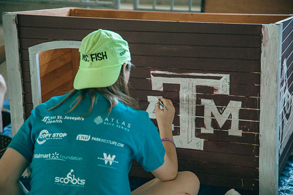 A student in a bright green MSC FISH hat, painting a dog house during Kyle Field Day.